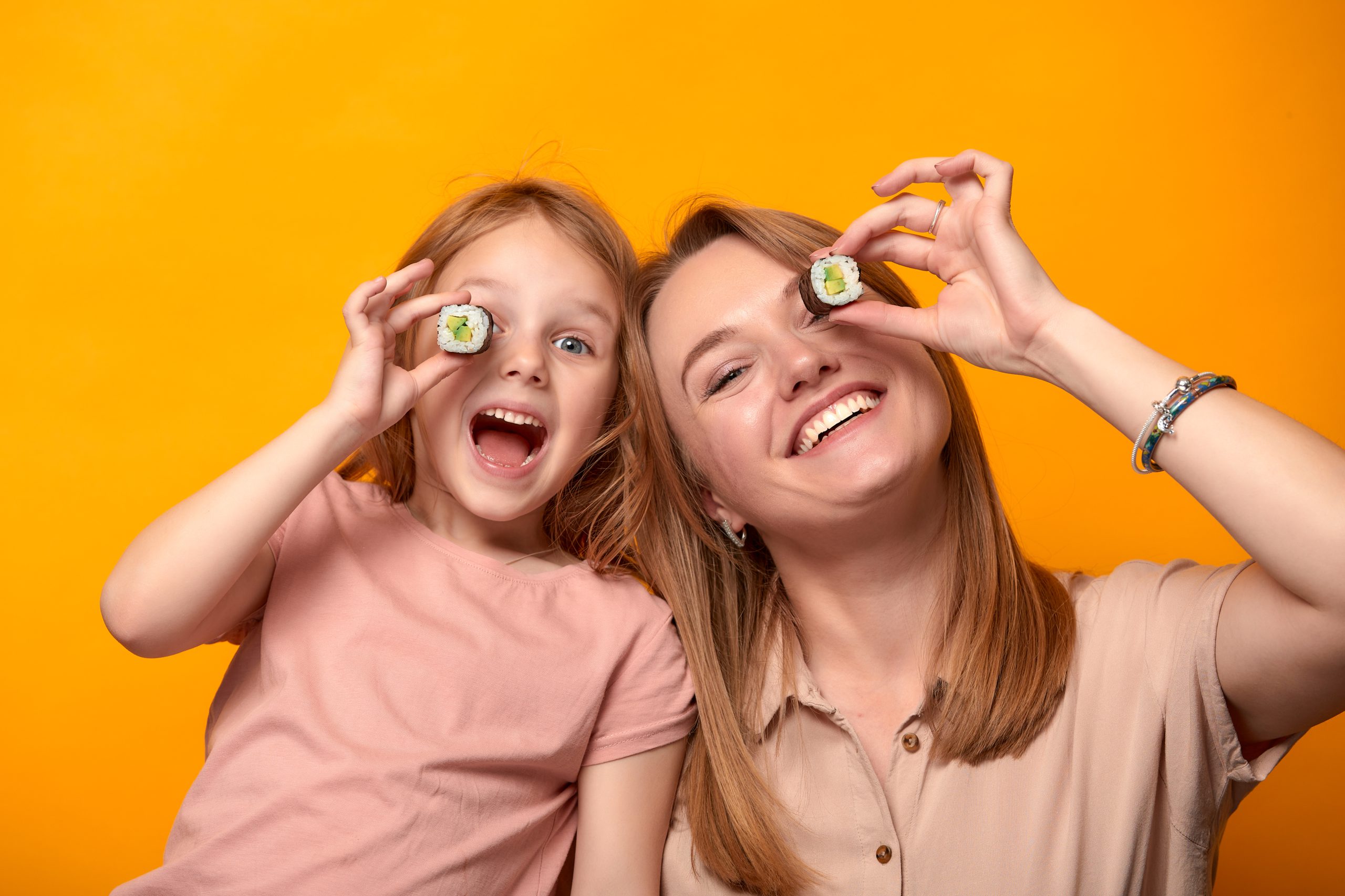 Funny mom with daughter holding sushi rolls in front of eyes on yellow background